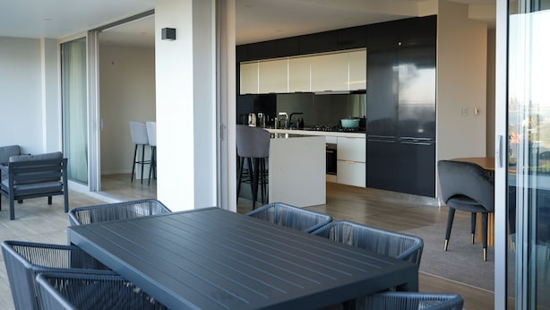 Una cocina moderna con armarios oscuros y una isla central, un comedor con mesa y sillas oscuras, y una vista a través de un ventanal.