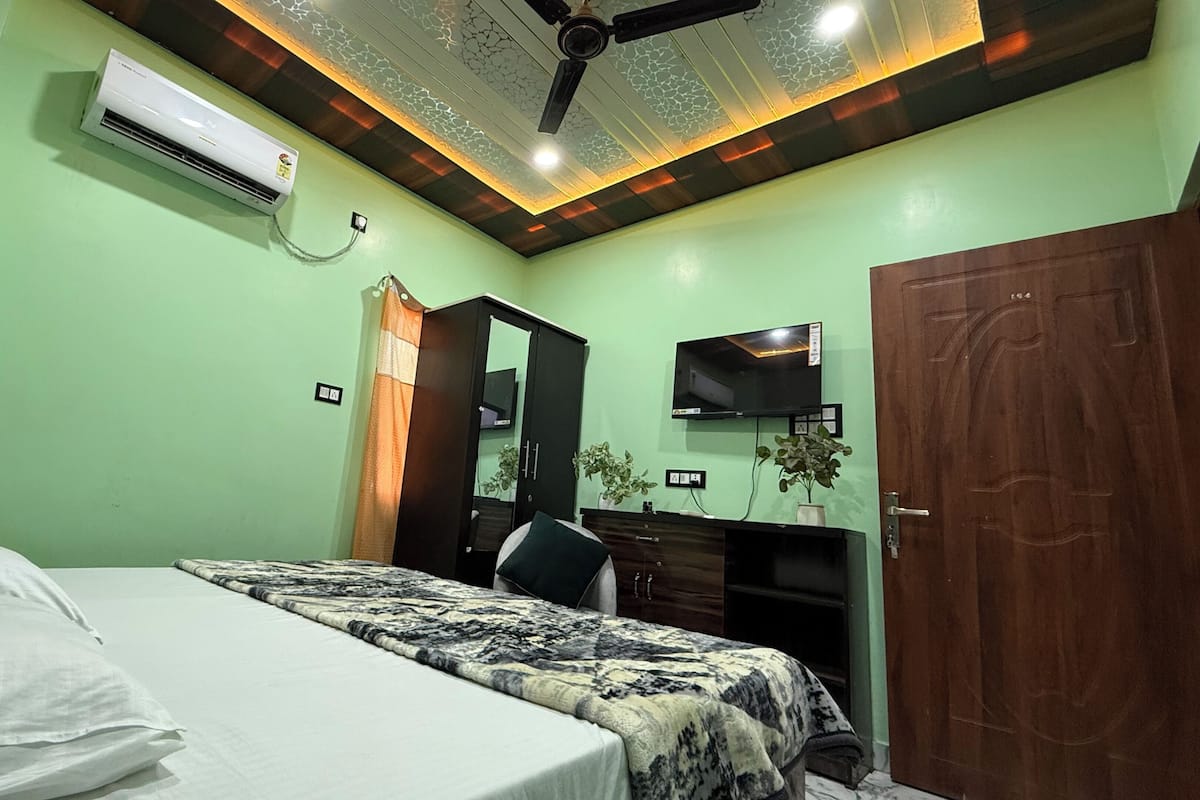A bedroom with a bed, a desk, a television, and an air conditioning unit.