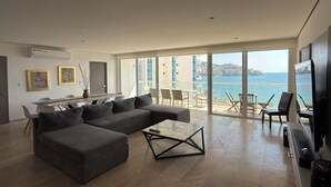 Ein modernes Wohnzimmer mit einer großen, grauen Eckcouch, einem Glastisch und Meerblick durch große Fenster.
