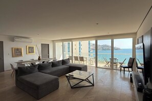 Ein modernes Wohnzimmer mit einer großen, grauen Eckcouch, einem Glastisch und Meerblick durch große Fenster.