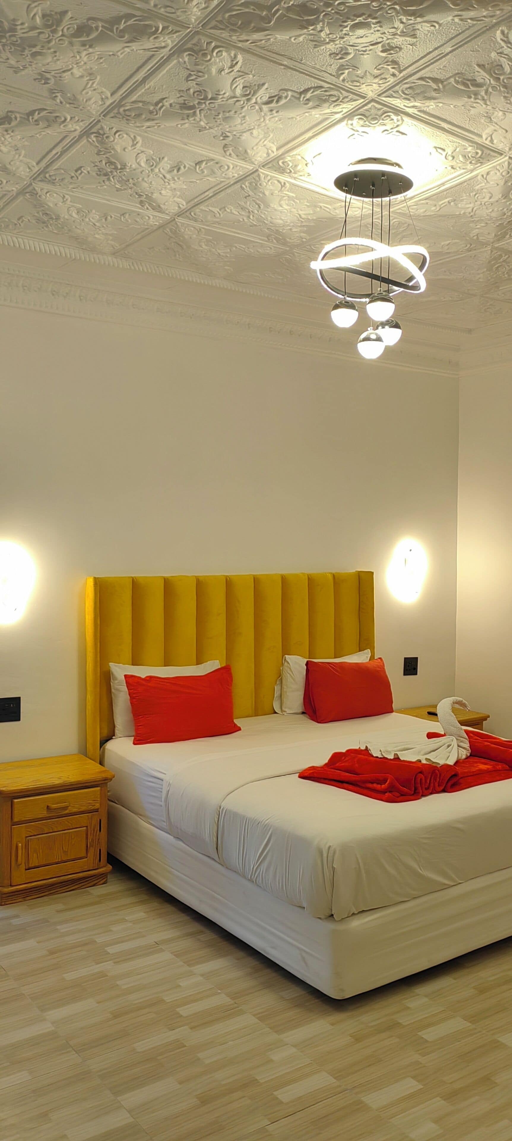 Une chambre d’hôtel moderne avec un grand lit, une table de chevet en bois, une lampe fixée au mur et un plafond décoratif.