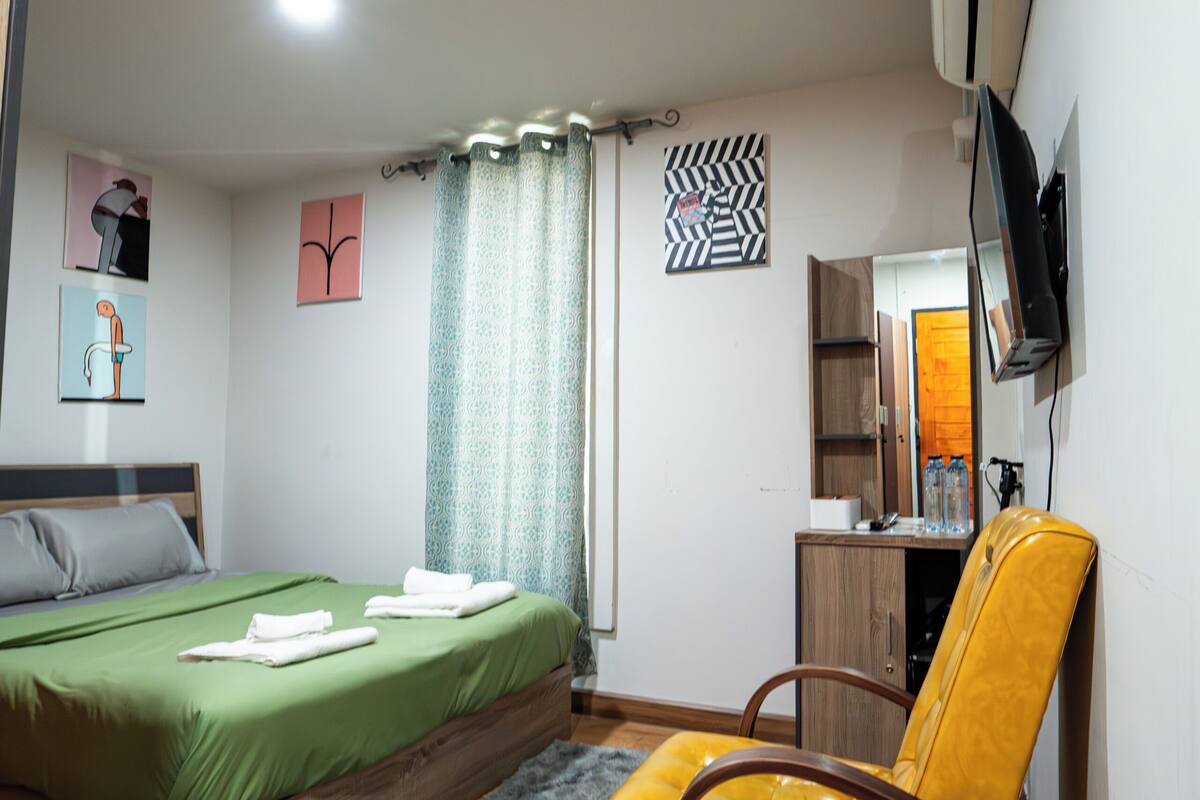 A bedroom with a bed, a chair, a desk, a mirror, and a television.