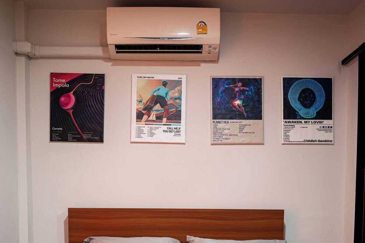 A hotel room with a bed, pillows, a wall-mounted air conditioner, and four posters on the wall.