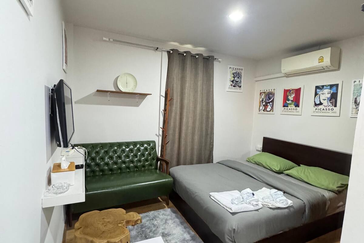 A compact hotel room with a green sofa, a single bed with green pillows, a white table, and a TV on a stand.