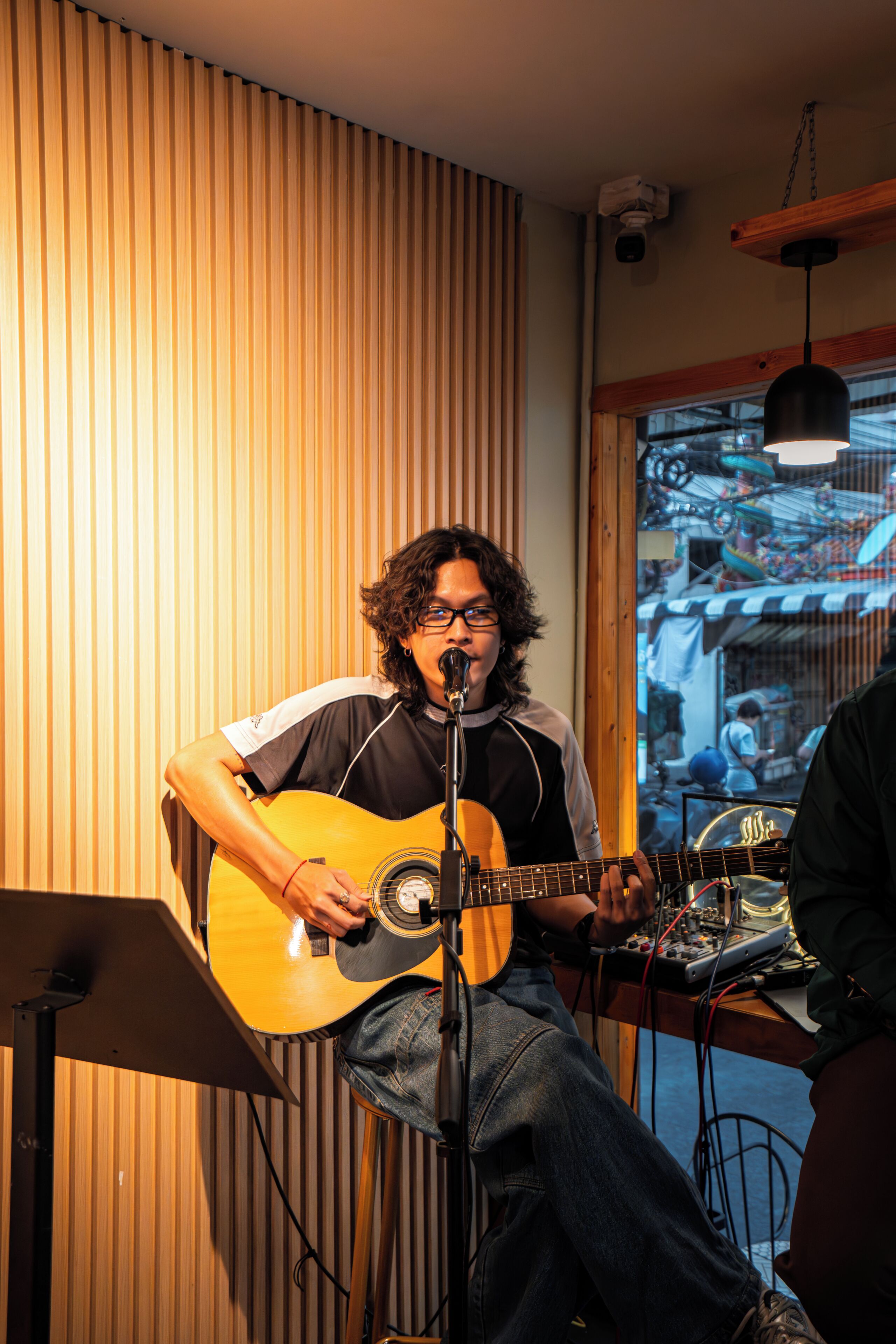 A person playing an acoustic guitar in a room with vertical wooden slats on the wall and a window with a view of a street scene.