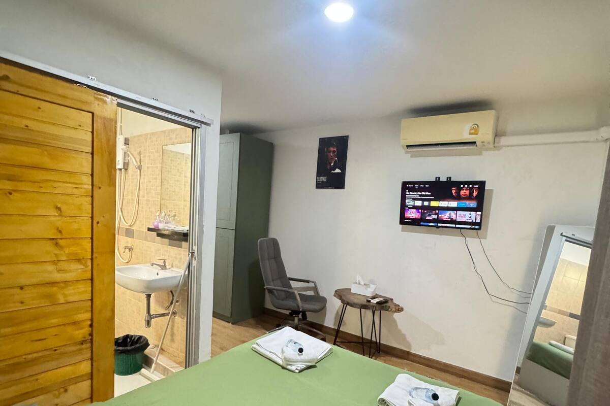 A room with a bed, a chair, a small table, a TV, and a bathroom visible through an open door.