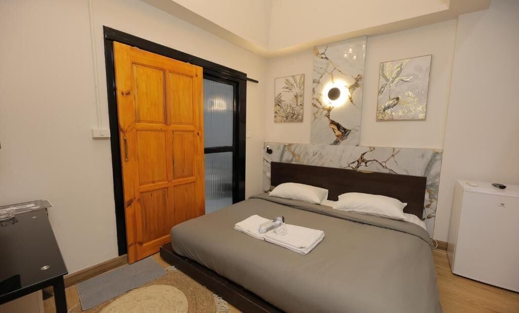 A bedroom with a bed, a wooden door, a small table, and a refrigerator.