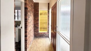 A narrow hallway with a brick wall, a wooden door, and a window with frosted glass. - Cosy 3BR Cottage Home w/ Ample Parking (Slacks Creek)
