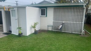 A small, single-story building with a green roof, a door, a window, and a patio area with a chair and a table. - Most Affordable Guesthouse in Hamilton? (Hamilton)