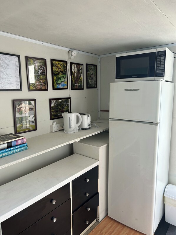 A compact kitchen with a refrigerator, microwave, kettle, and coffee maker. There are framed pictures on the wall and a small shelf with books.