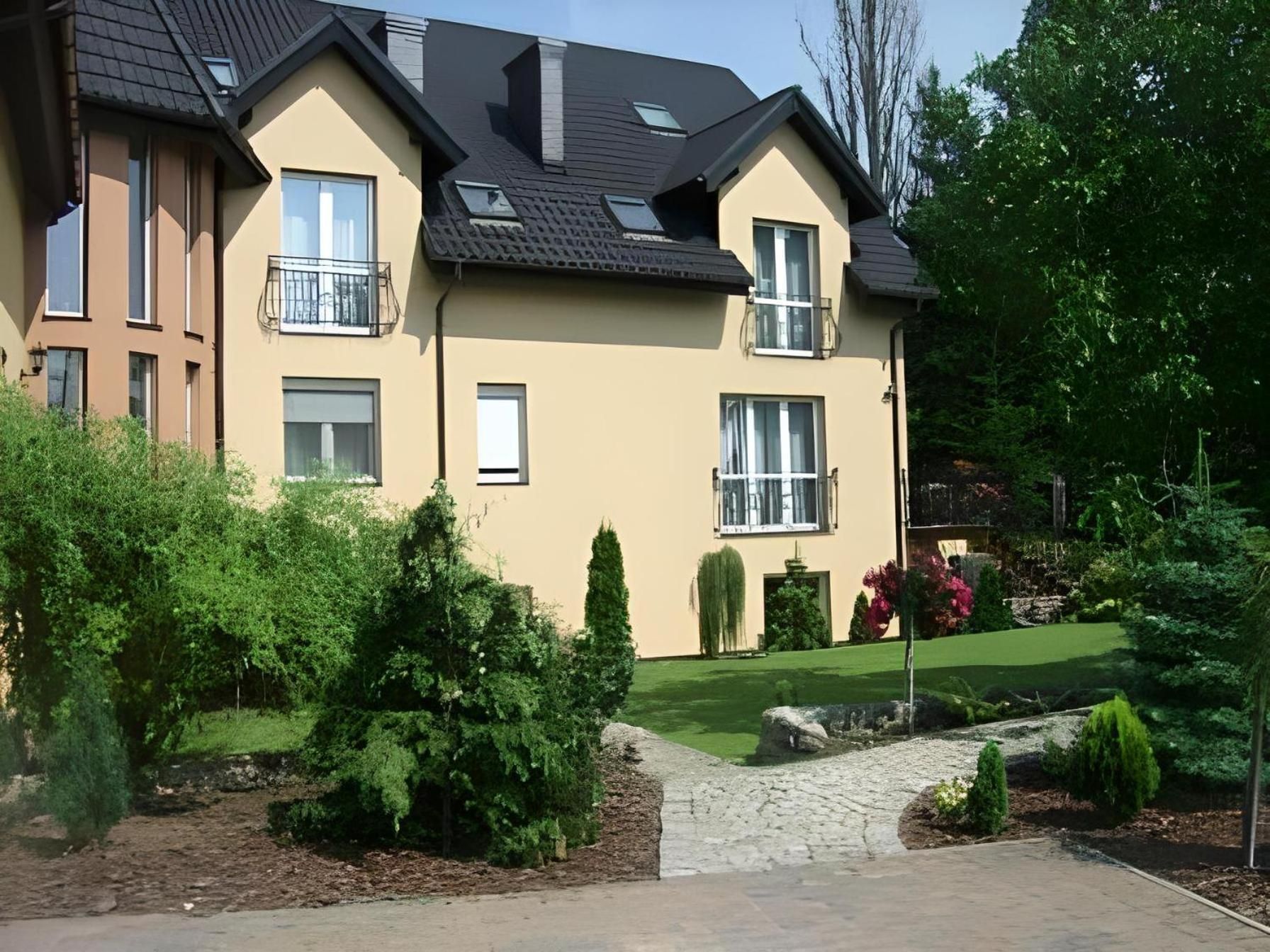A two-story residential building with a sloped roof, multiple windows, and a well-maintained garden with various shrubs and a lawn.