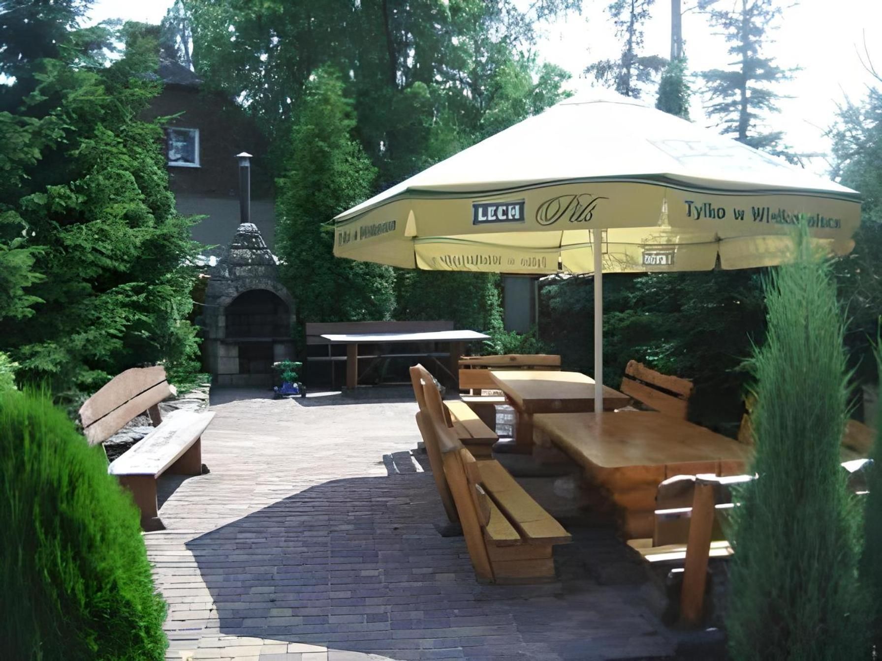 An outdoor seating area with wooden tables and benches, an umbrella, and a stone structure in the background.