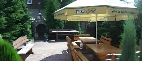 An outdoor seating area with wooden tables and benches, an umbrella, and a stone structure in the background.