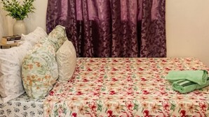 A bedroom with a bed, floral bedspread, pillows, a potted plant, and purple curtains.
