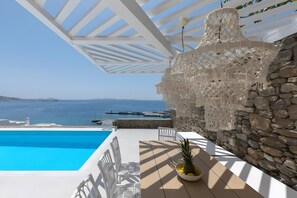 Un espace aménagé au bord de la piscine, avec vue sur la mer, doté de mobilier blanc et d’un mur en pierre.
