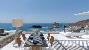 A dining table set with blue plates and glasses, overlooking a marina with boats and a cruise ship, with a clear blue sky above. - Dream Baby with Private Pool (Mykonos)