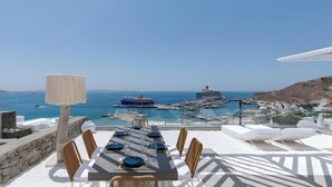 Ein Esstisch ist gedeckt mit blauen Tellern und Gläsern, mit Blick auf einen Yachthafen mit Booten und einem Kreuzfahrtschiff, über dem ein klarer, blauer Himmel strahlt.