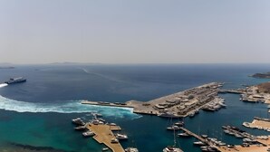 Ein Küstenhafen mit vertäuten Booten, einer Mole, die ins Wasser ragt, und einem Schiff in der Ferne.
