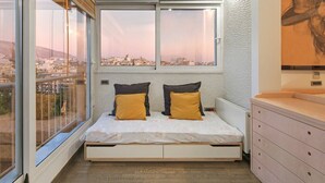 Quarto com cama, duas almofadas, penteadeira e uma grande janela com vista para a cidade.