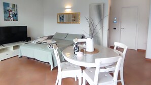 Una sala moderna con televisor de pantalla plana, una mesa de comedor con sillas blancas, un sofá, un espejo grande y un aplique de techo.