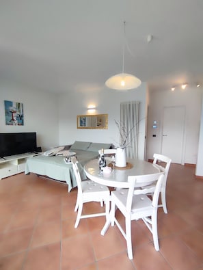Ein modernes Wohnzimmer mit einem Flachbildfernseher, einem Esstisch mit weißen Stühlen, einem Sofa, einem großen Spiegel und einer Deckenleuchte.
