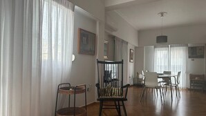 A spacious living room with a wooden floor, a rocking chair, a dining table with chairs, and a large window with sheer curtains. - AthensView Skyline: 5th fl, 2BR, Acropolis View (Athina)