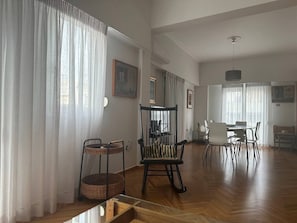 Ein geräumiges Wohnzimmer mit Holzboden, einem Schaukelstuhl, einem Esstisch mit Stühlen und einem großen Fenster mit durchsichtigen Vorhängen.