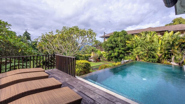 A pool with a wooden deck and lounge chairs, surrounded by lush greenery and a building in the background. - O'hare Villa  (Jimbaran)