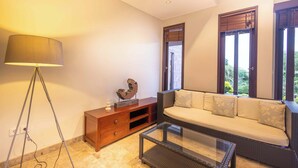 A modern living room with a sofa, a glass coffee table, a lamp, a wooden cabinet, and a window with blinds. - O'hare Villa  (Jimbaran)