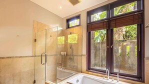 A bathroom with a glass-enclosed shower, a bathtub, and a window with a view of greenery. - O'hare Villa  (Jimbaran)