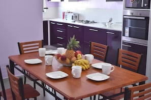 Ein Esstisch mit Tellern, Tassen und einer Obstschale in einer Küche mit violetten Schränken und modernen Geräten.
