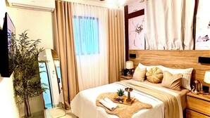 Ein Hotelzimmer mit Bett, Deckenventilator, Nachttisch, Lampe, Spiegel, Topfpflanze und einem Fenster mit Vorhängen.