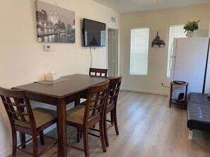 Ein Essbereich mit Holztisch und Stühlen, einem an der Wand befestigten Flachbildfernseher und einem Kühlschrank mit Pflanze darauf.