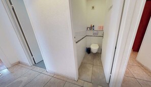 A narrow hallway leading to a bathroom with a toilet, a small shelf with toiletries, and a tiled floor. - Private Room in a Shared Flat - Lyon 8 (Lyon)