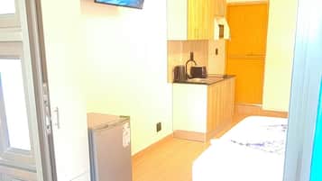 A compact room with a bed, a small kitchenette, and a television mounted on the wall.