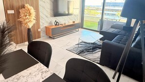 A modern living room with a flat-screen TV, a wooden accent wall, a large window with a view, and a black sofa. - Apartment TM lagoons Village 141 (Torrevieja)