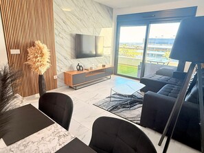 A modern living room with a flat-screen TV, a wooden accent wall, a large window with a view, and a black sofa. - Apartment TM lagoons Village 141 (Torrevieja)