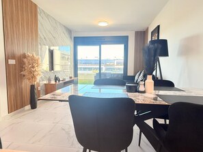 A modern living room with a large window, a flat-screen TV, a dining area with chairs, and a view of the cityscape. - Apartment TM lagoons Village 141 (Torrevieja)