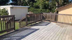 A wooden deck with a railing, surrounded by greenery and a building.