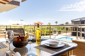 A dining table with a fruit bowl, wine glasses, and a bottle, overlooking a swimming pool and a building with a red roof. - Sea View & Luxury Apartment Domania' with Wi-Fi and Air Conditioning (Καληκρατειας Σωζόπολη)