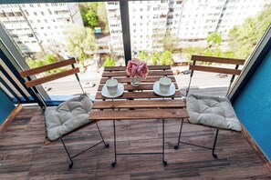 Ein Balkon mit Holztisch, zwei gepolsterten Stühlen, einer Blumenvase und Blick auf eine Wohngegend.