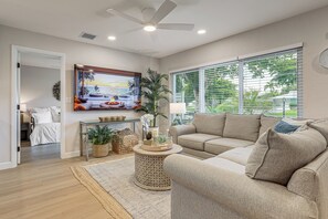 Ein Wohnzimmer mit einem beigefarbenen Sofa, einem runden Couchtisch, einem Gemälde und einem großen Fenster mit Jalousien.