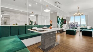 A modern hotel room with a green sofa, a dining area, a large window with a city view, and a chandelier. - Narel by Kozystay - Kuningan (Jakarta)
