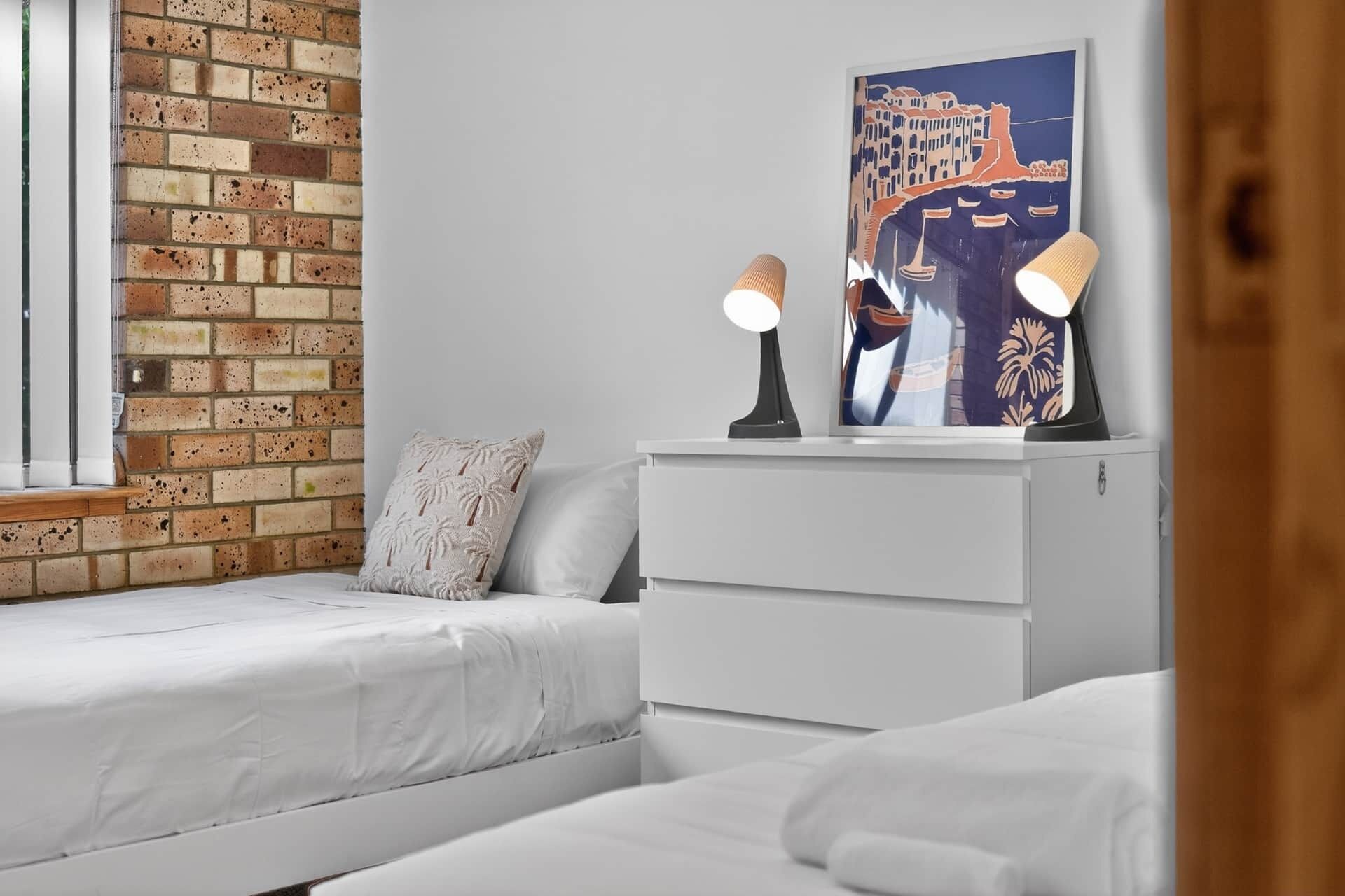 A bedroom with two beds, a brick wall, a white dresser, and a painting of a coastal scene.