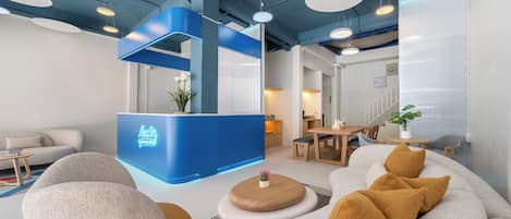 Un área de recepción moderna con un mostrador azul, asientos con cojines e iluminación colgante.