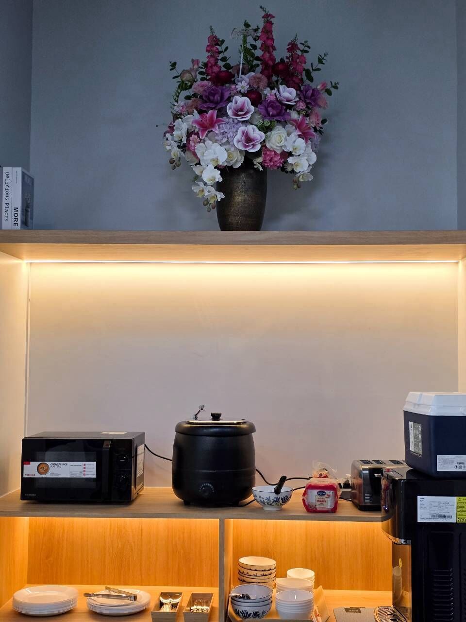 Una cocina con microondas, hervidor y tostadora sobre la encimera, un florero con flores en la pared, y vajilla y utensilios ordenados abajo.