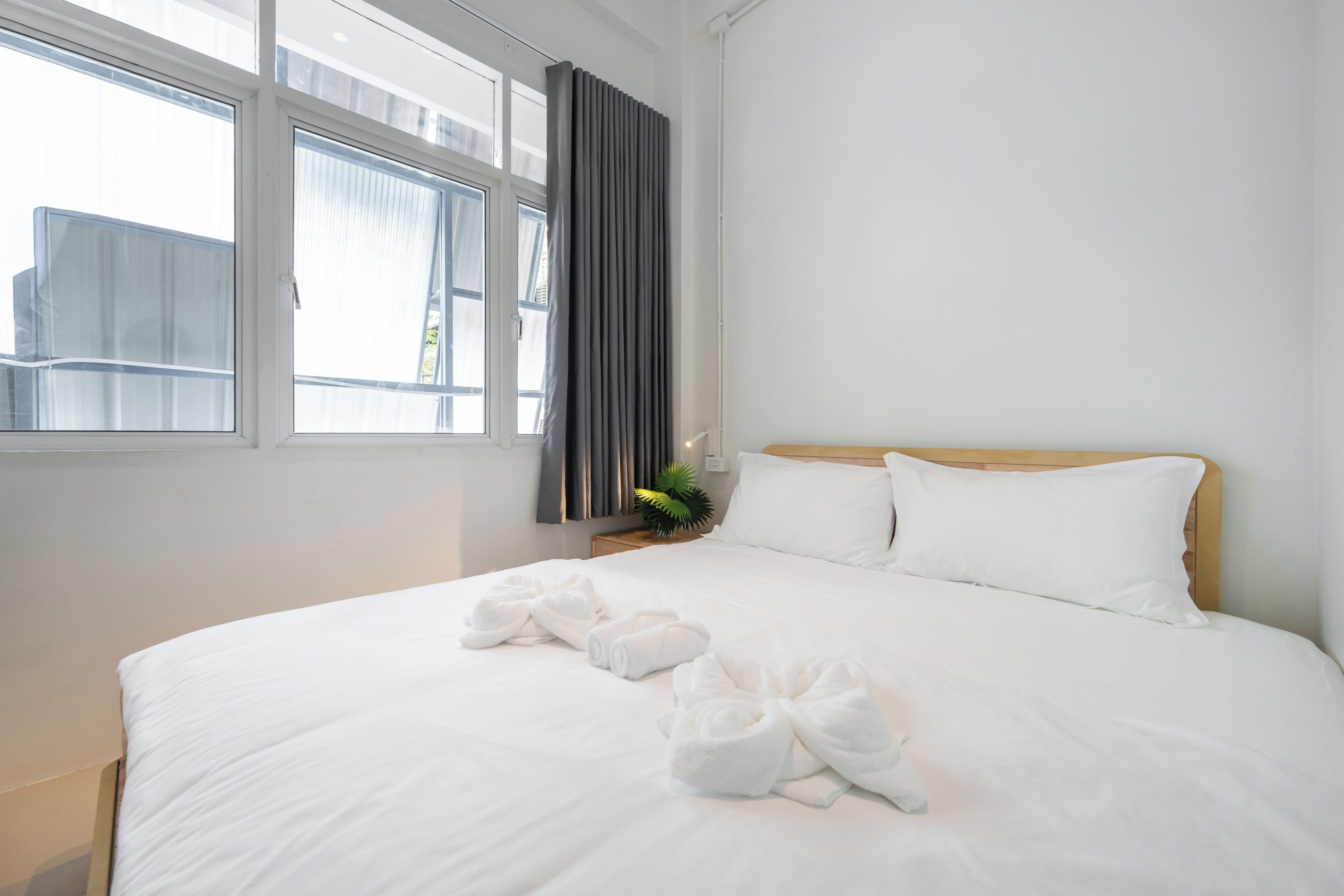 Una camera d'albergo con un letto grande, lenzuola bianche, una finestra con tende e una piccola pianta sul comodino.