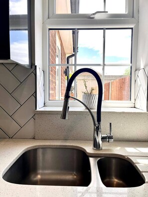 A kitchen with a double stainless steel sink, a modern faucet, and a window with a view of a brick building and a wooden fence.