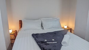 A neatly made bed with white linens, two pillows, a dark blanket, and a rolled towel. There are two bedside lamps on wooden nightstands. - The Hub Train House Amazing Gents code (Zanzibar)