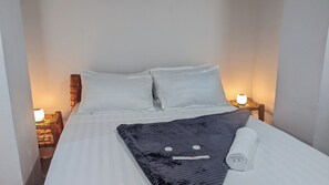 Uma cama bem arrumada com lençóis e almofadas brancas, um cobertor cinza escuro e uma toalha enrolada ao lado. Existem duas luminárias de cabeceira em mesas de cabeceira de madeira.
